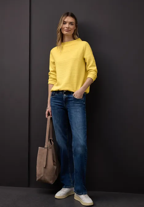 Frau in gelbem Pullover, blauen Jeans und weißen Sneakern mit brauner Tasche vor schwarzem Hintergrund.